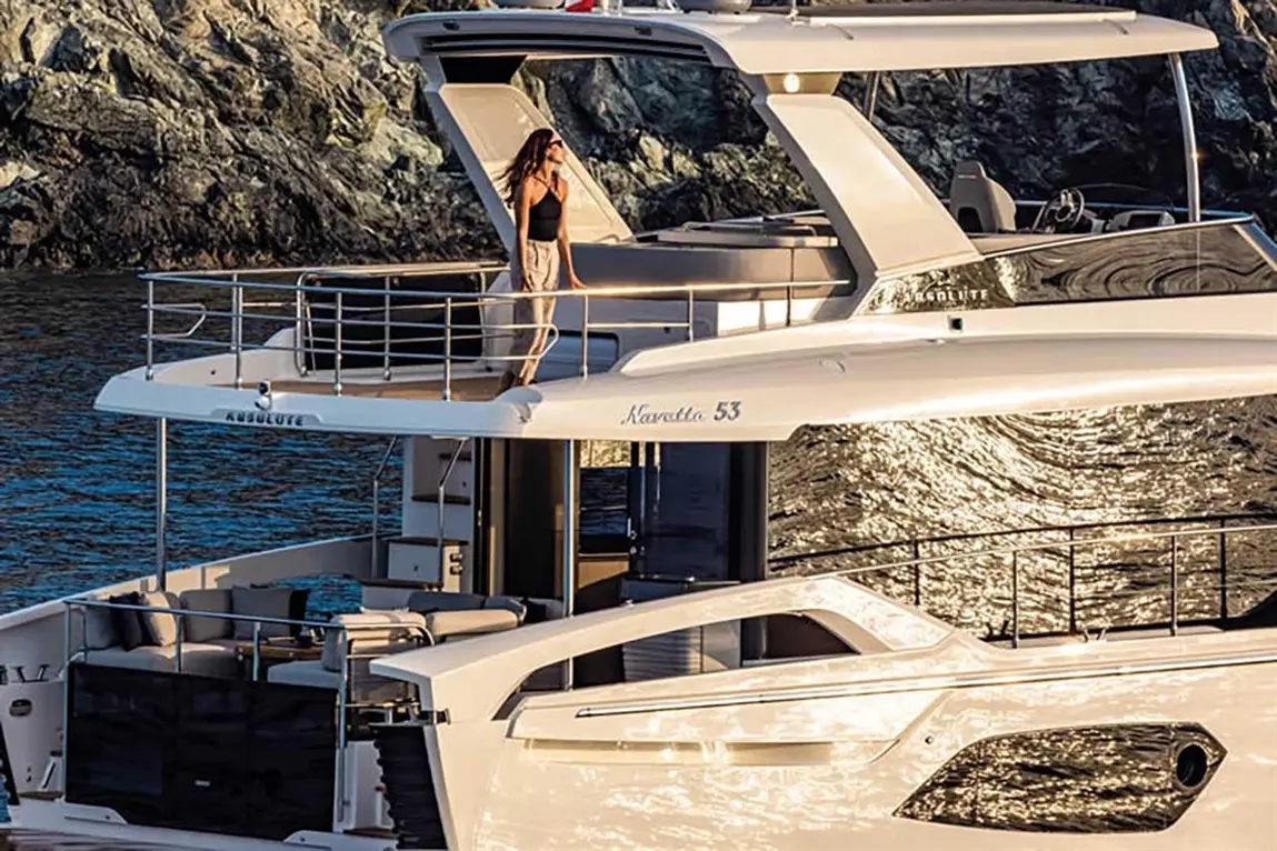 A woman stands on the deck of an Absolute Navetta 53 yacht, with shimmering water and rocky cliffs in the background.